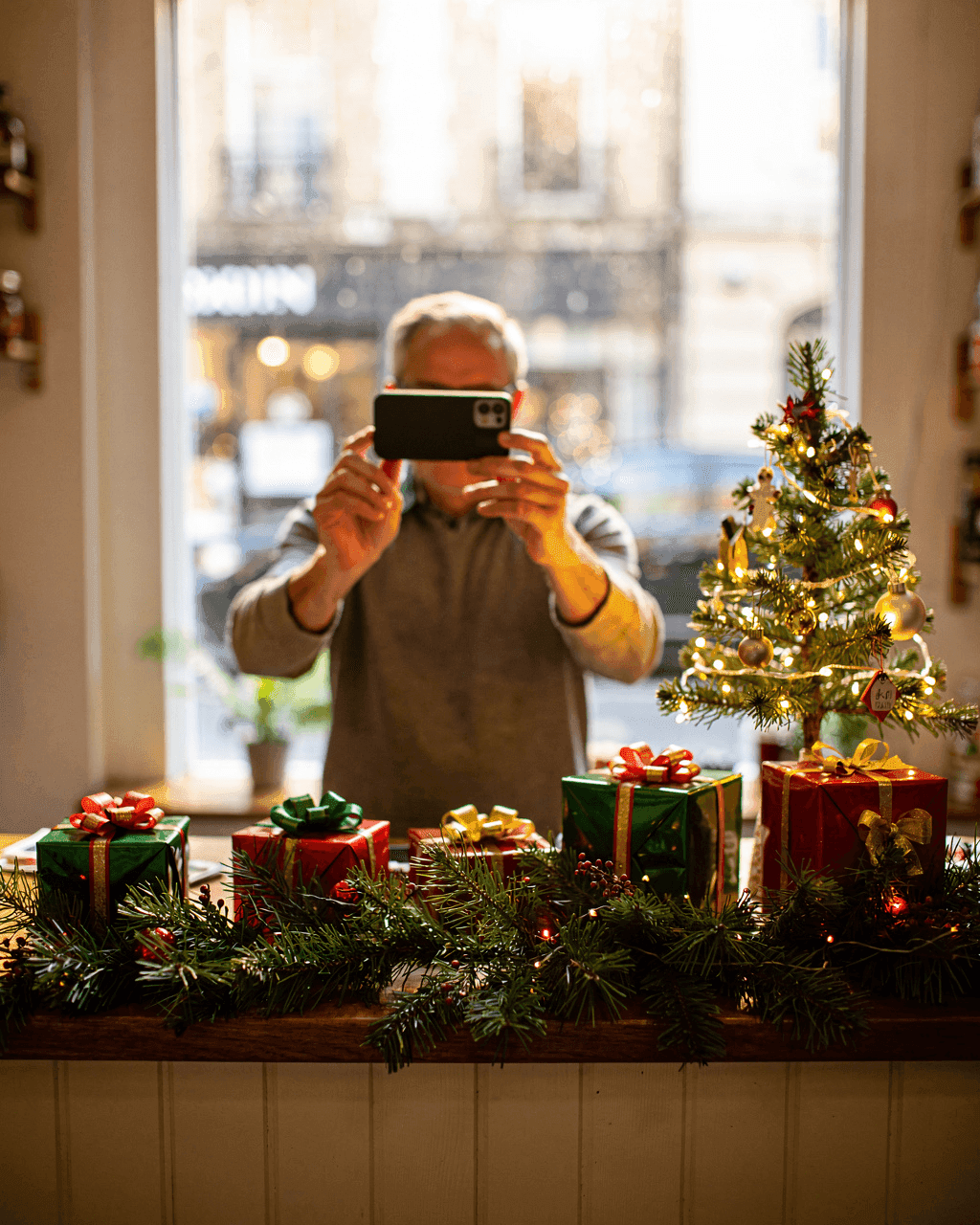 Shop owner photographing a Christmas-decorated shop counter with a phone, a small lit tree and wrapped gifts on the counter, daylight through the storefront window