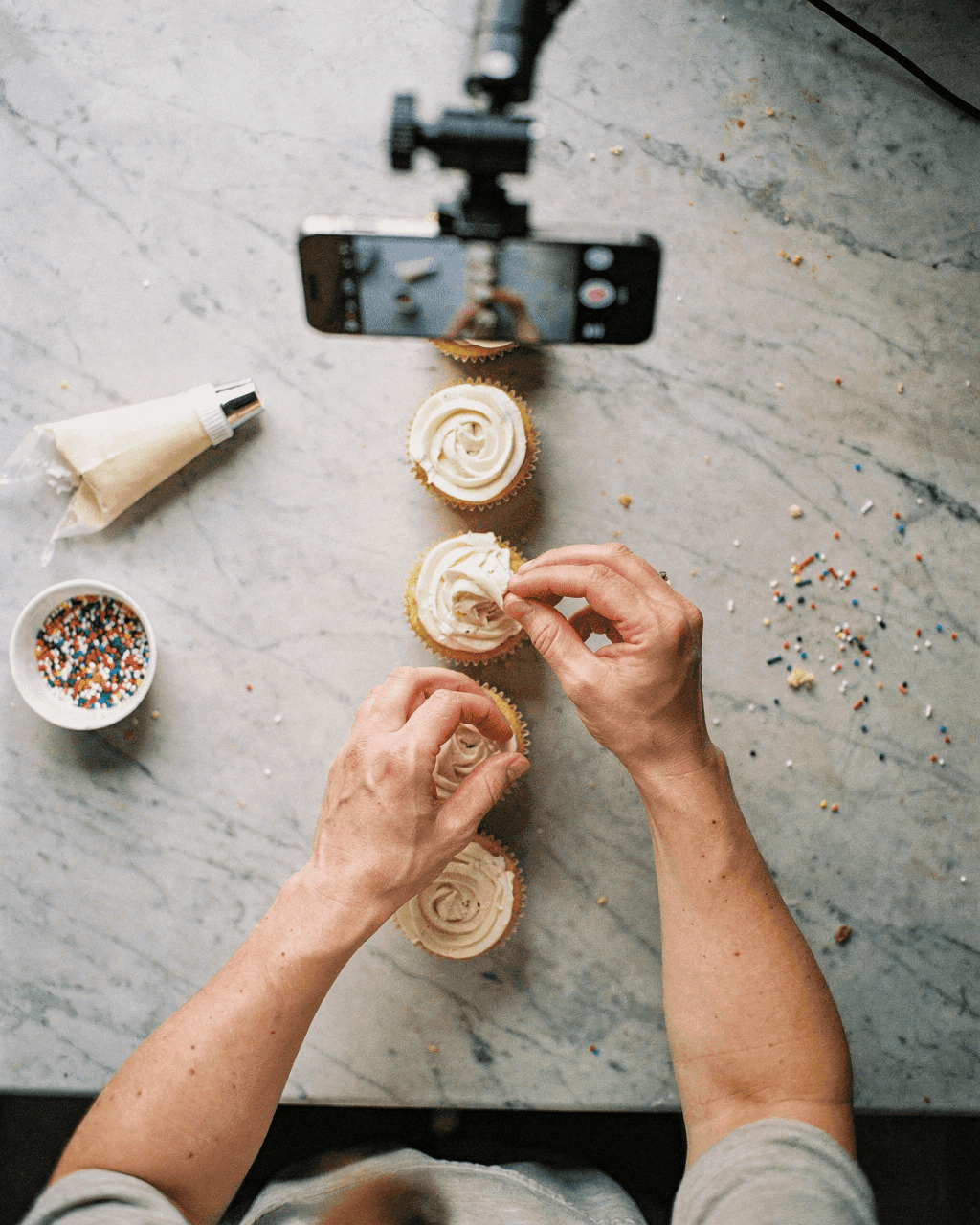 Top-down view of hands frosting cupcakes while a phone films from above