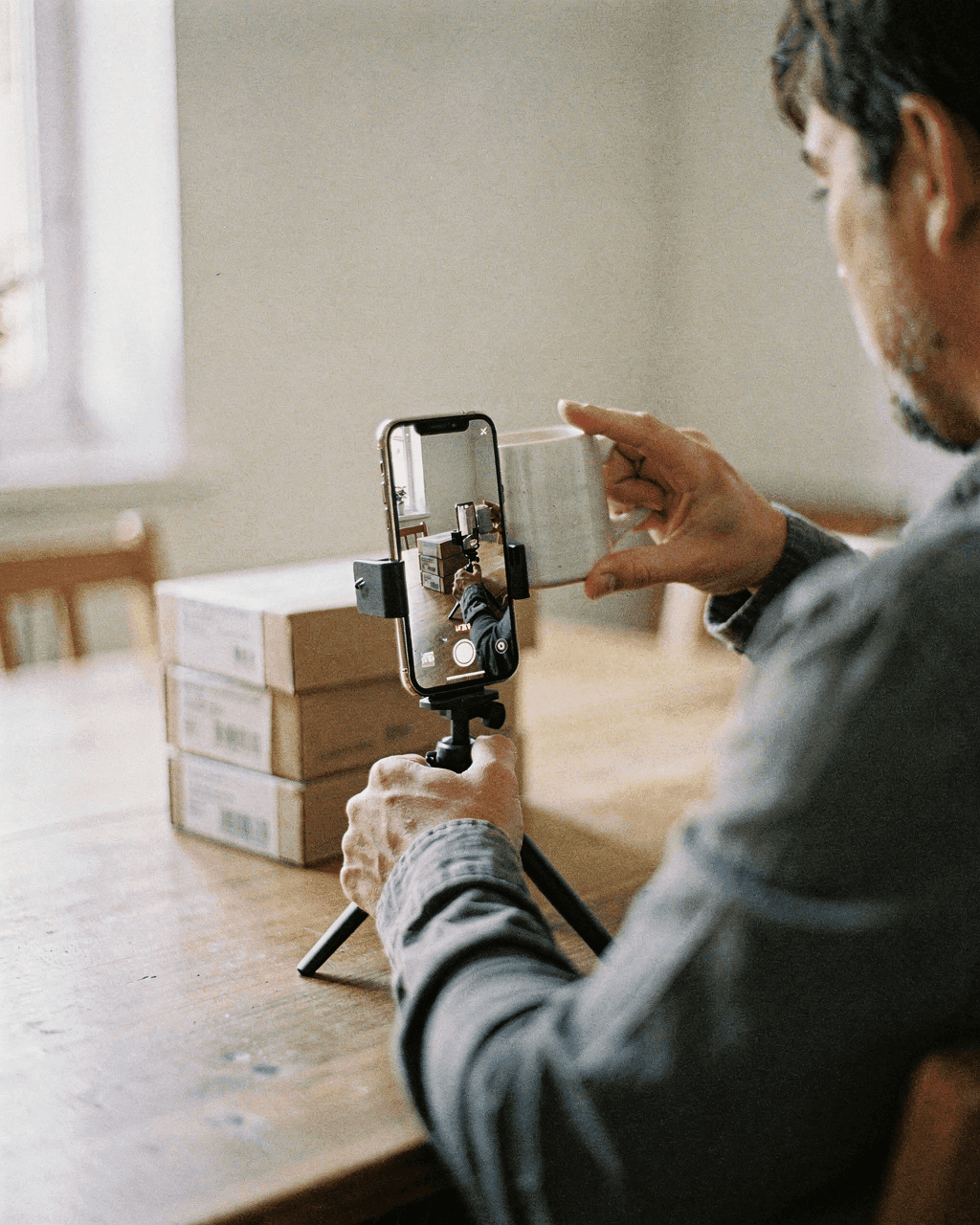 Seller seen from behind holding a grey ceramic mug in front of a phone on a tripod, stacked shipping boxes on the table