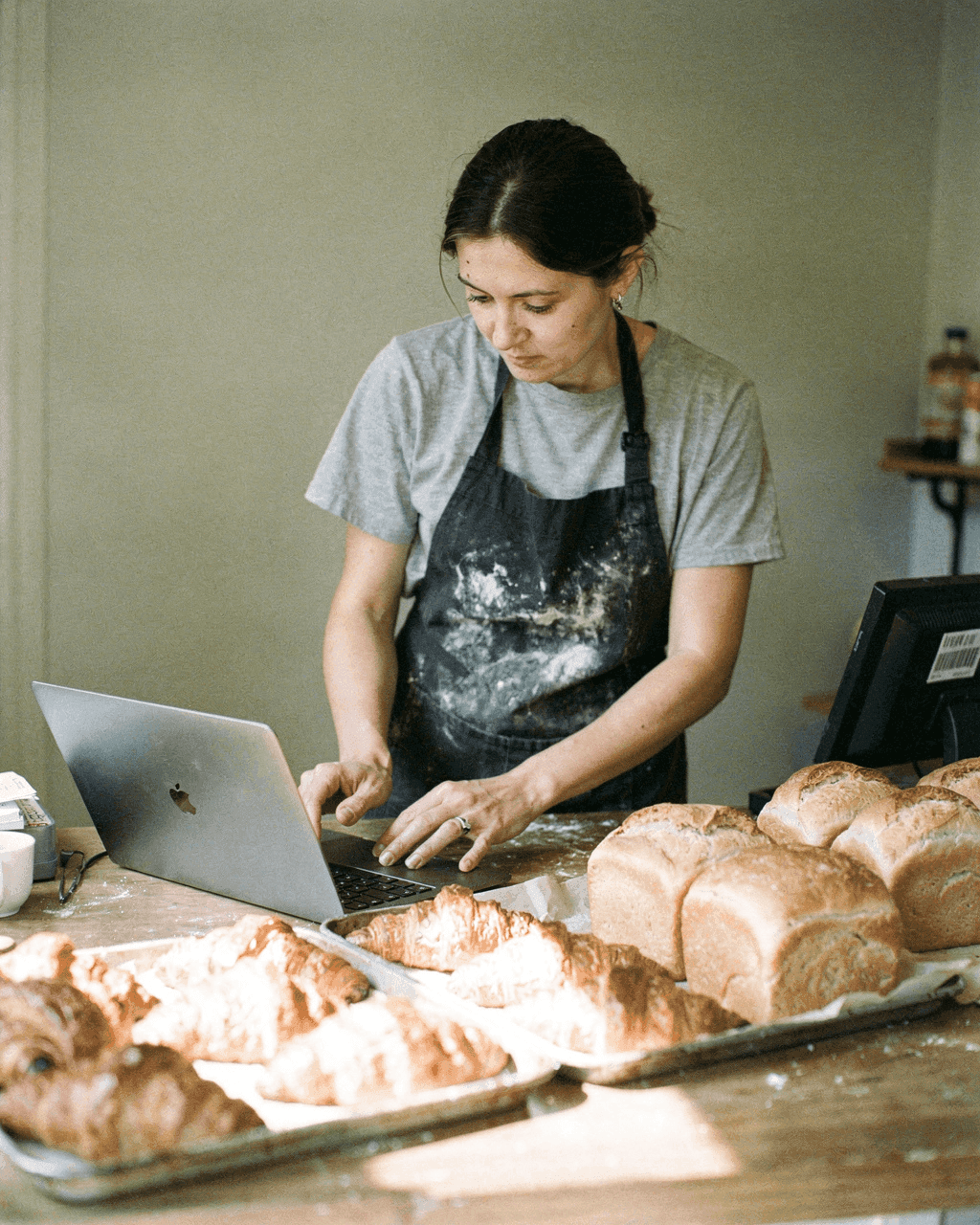 Bakery owner choosing photos on a laptop behind her counter, fresh bread beside her