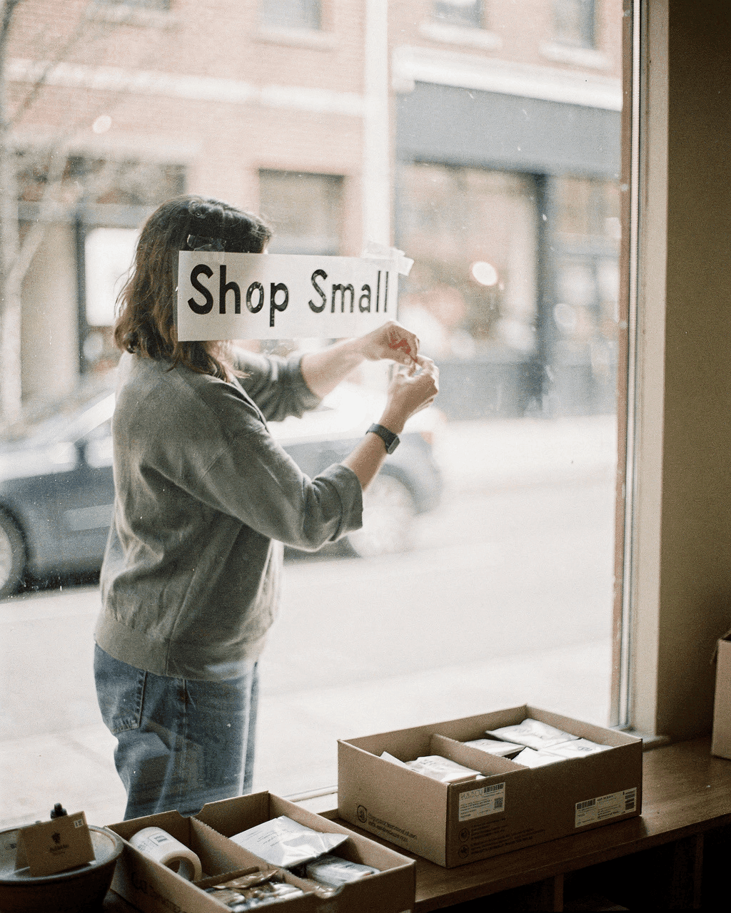 Shop owner taping a handwritten Shop Small sign to the inside of their storefront window, boxes of inventory nearby